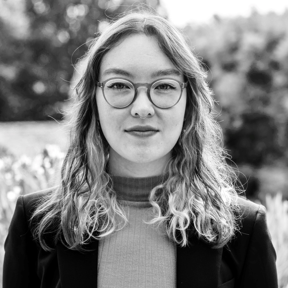 Black and white portrait of Lena Hänni, female, fair-skinned person with a turtleneck sweater, blazer, glasses, and long, curly hair