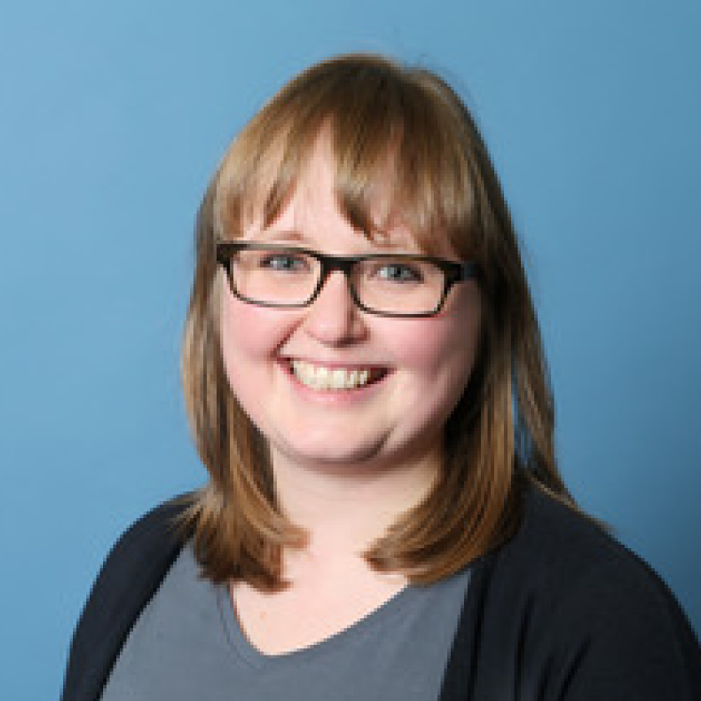 Portrait of Dr. Camilla Bernardasci, a female, fair-skinned person wearing a blue-gray top, a black jacket over it, glasses, and shoulder-length brown hair.