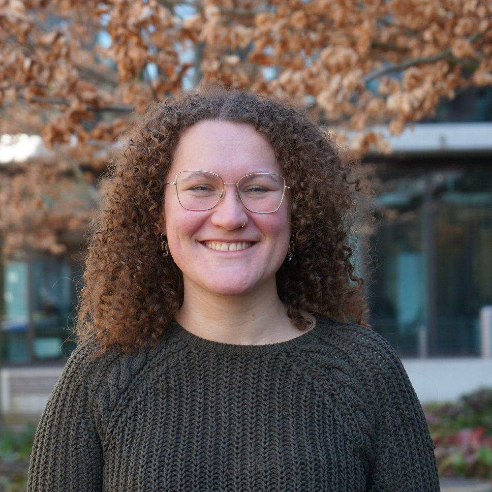 Portrait Ladina Bischof, weiblich lesbare, hellhäutige Person mit grauem Strickpullover, Brille und schulterlangen, lockigen, braunen Haaren