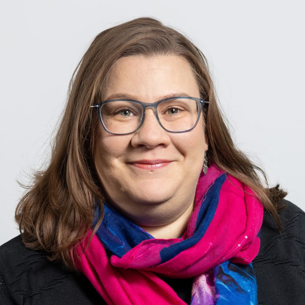 Portrait of Martina Schweizer, female, fair-skinned person with a black top, pink-blue scarf, glasses, and shoulder-length, brown hair